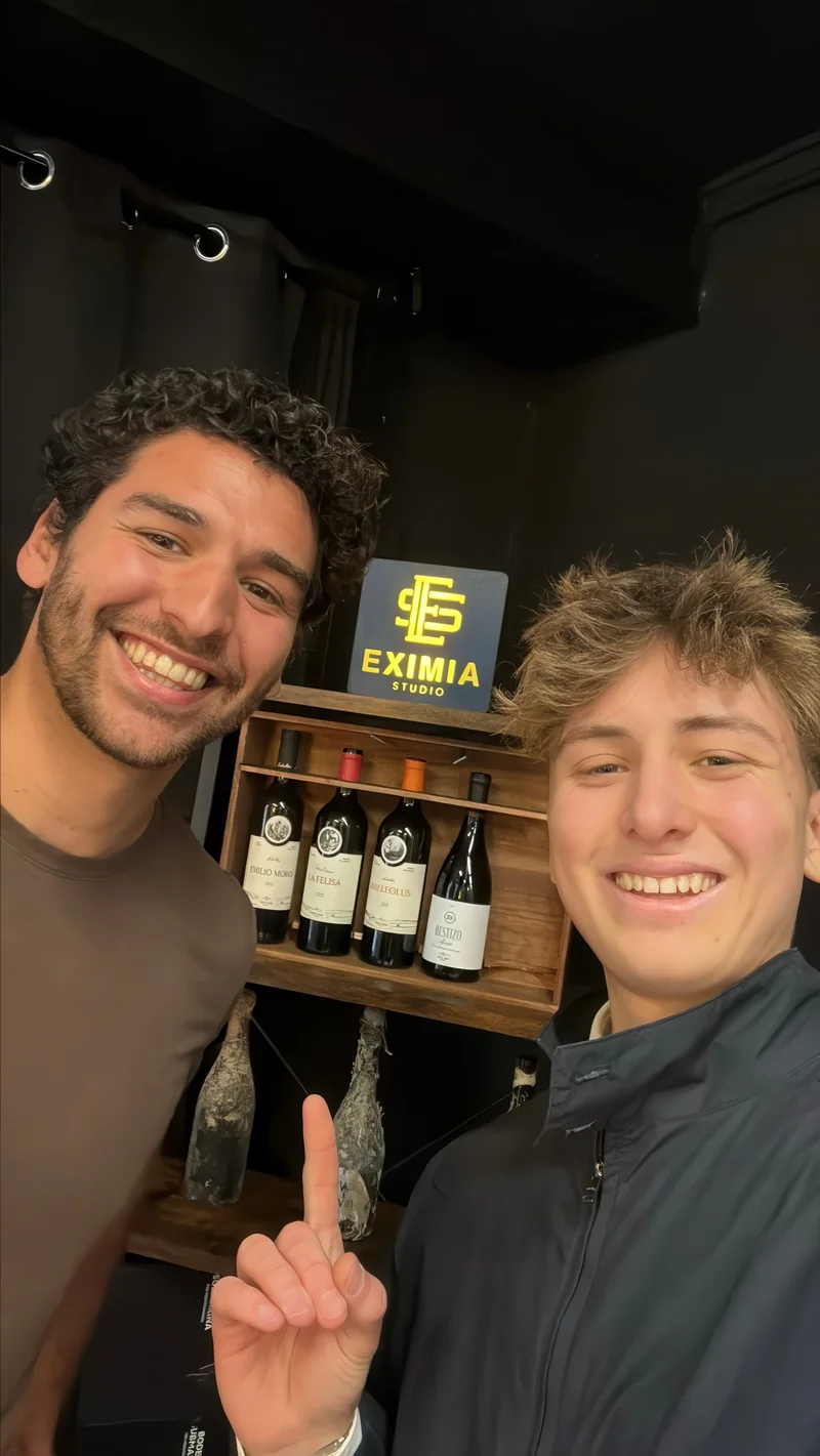 Uriel y un compañero sonriendo delante del rótulo dorado de Eximia Studio, con un botellero de vinos detrás.