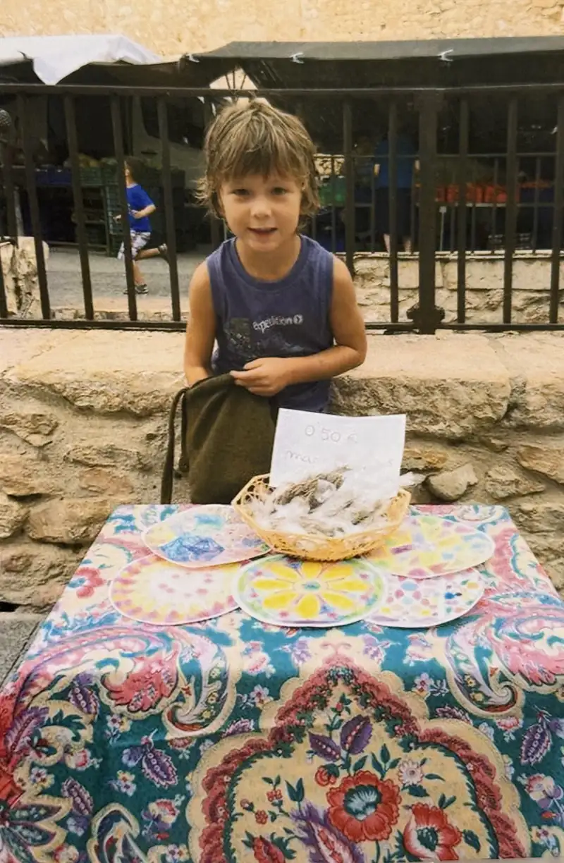 Uriel de niño, detrás de una mesa cubierta con un mantel de flores, vendiendo mandalas de papel en la plaza del pueblo junto a un cartel de 0,50 €.