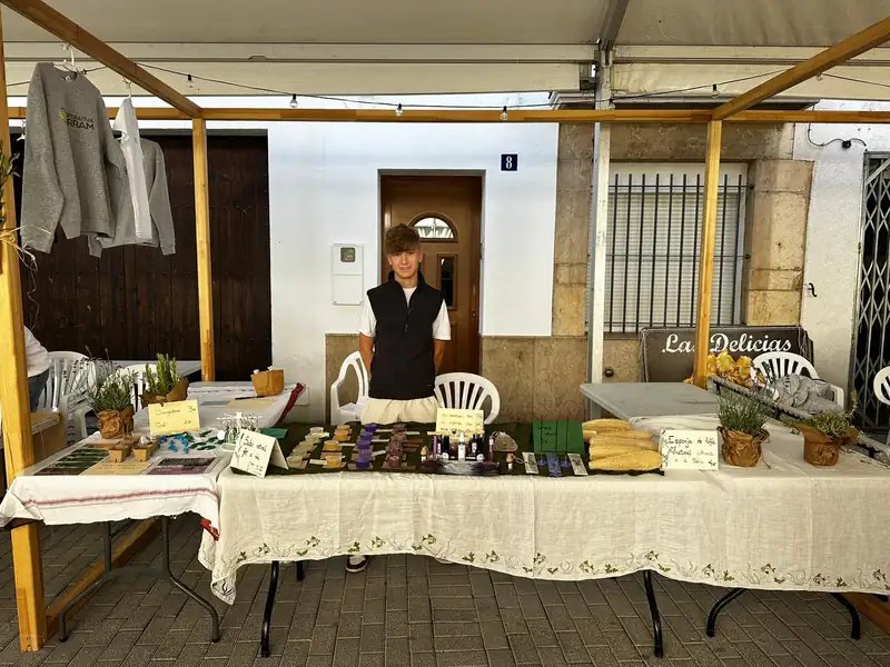 Uriel tras dos mesas con mantel bordado en una calle del pueblo, con los jabones en fila y el rótulo «Las Delicias» en la pared del fondo.