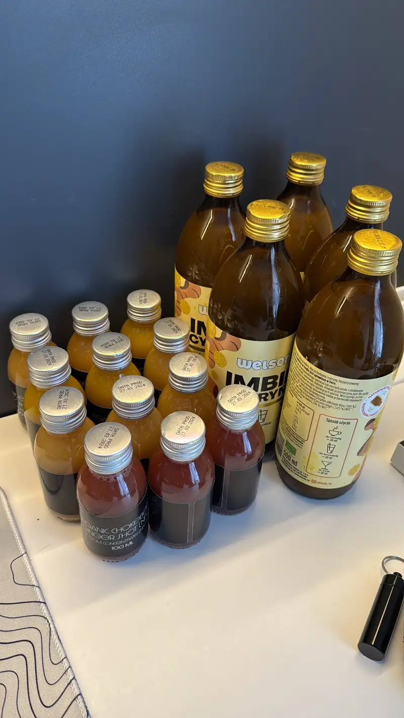 Botellas de cristal ámbar con tapón dorado y una hilera de shots pequeños de jengibre sobre una mesa blanca.