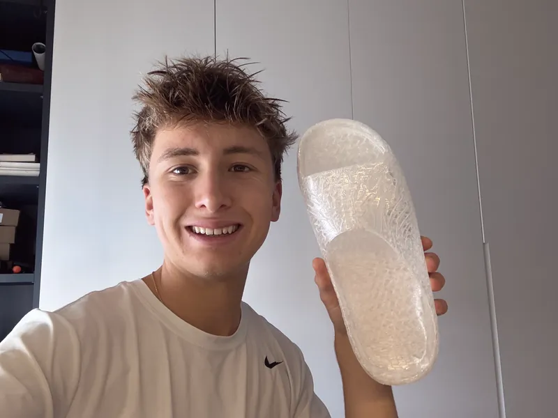 Uriel sonriendo mientras sostiene en alto una chancla translúcida impresa en 3D.